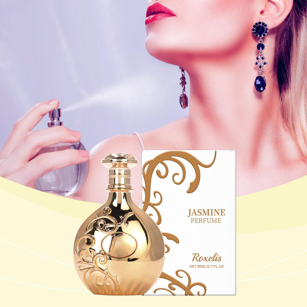 A perfume bottle and its box for Roxelis Jasmine Perfume, shown in front of a model spraying the fragrance. The perfume bottle is metallic gold with ornate scrollwork, and the box is white with gold decorative flourishes and text.